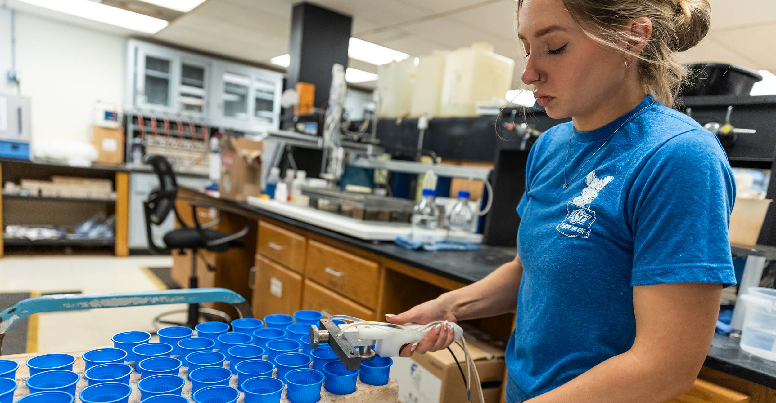 A female laboratory technician in a blue shirt uses a hand-held dispensing pump tool to add water to soil samples in small blue cups lined up in a styrofoam tray.