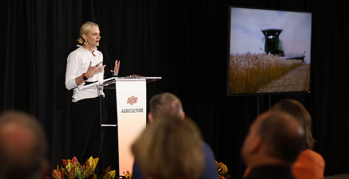 Kayse Shrum stands at a podium with the logo OSU Agriculture. She is wearing a white shirt and black pants. Her hair is slicked back in a bun. A screen in the background displays a photo of a John Deere combine harvesting wheat.