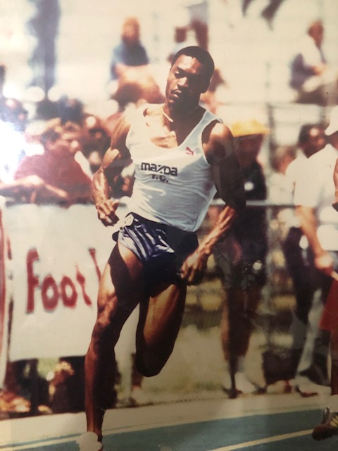 James Butler runs in a sprint on a track. He's wearing blue shorts, a white tank top and track shoes.