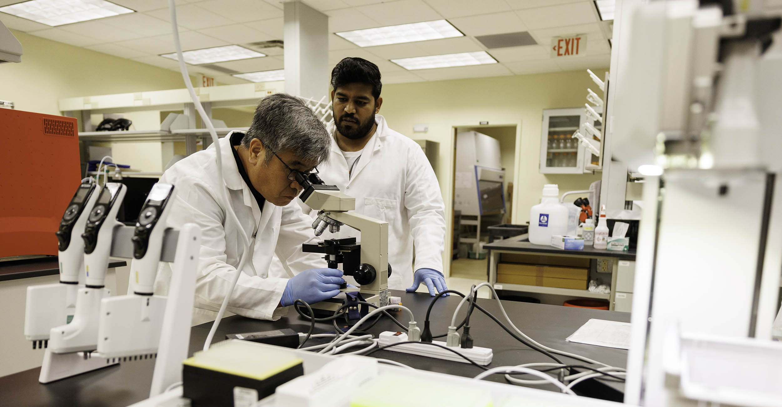 2024-onehealthlab-banner.jpg Hong Hwang looks through a microscope as Samuel Jeyasingh stands next to him in a laboratory.