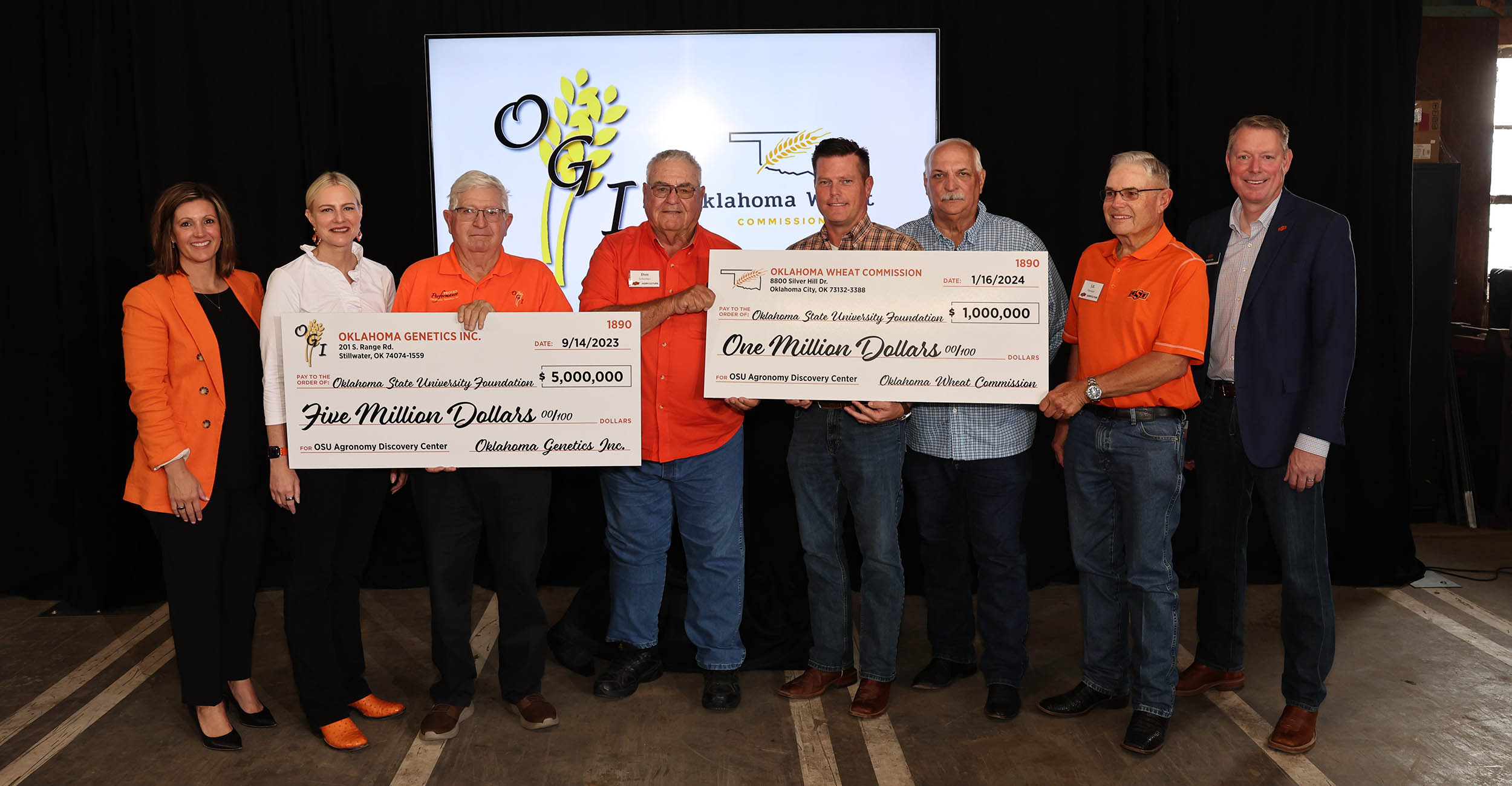 A group of six men and two women wearing orange, white or black shirts stand together holding two large photo prop checks in front of a black backdrop with a screen that displays the Oklahoma Genetics Inc. and Oklahoma Wheat Commission logos. A group of six men and two women wearing orange, white or black shirts stand together holding two large photo prop checks in front of a black backdrop with a screen that displays the Oklahoma Genetics Inc. and Oklahoma Wheat Commission logos.
